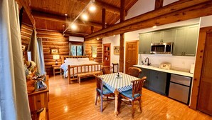 Interior de una cabaña de troncos con una mesa de comedor de madera, una cama y una zona de cocina con microondas y fregadero.