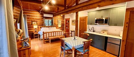 A log cabin interior with a wooden dining table, a bed, and a kitchen area with a microwave and sink.