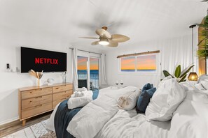Una moderna camera d'albergo con un letto, una cassettiera in legno, una ventola a soffitto, un televisore che mostra 'NETFLIX' e vista sul tramonto dalla finestra.