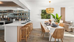 A modern kitchen with white cabinetry, a central island with bar stools, and a dining area with a white table and wicker chairs. - Brand New Luxury | Sunset Balcony | 3 Pools (panama city beach)