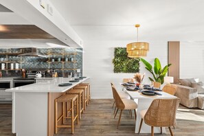 Ett modernt kök med vita skåp, en köksö med barstolar och en matplats med ett vitt bord och flätade stolar.