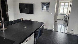 A modern, well-lit room with a dark wooden floor, a large black table with chairs, a flat-screen TV mounted on the wall, and a framed picture above the TV. - Apartment morteau center (Morteau)