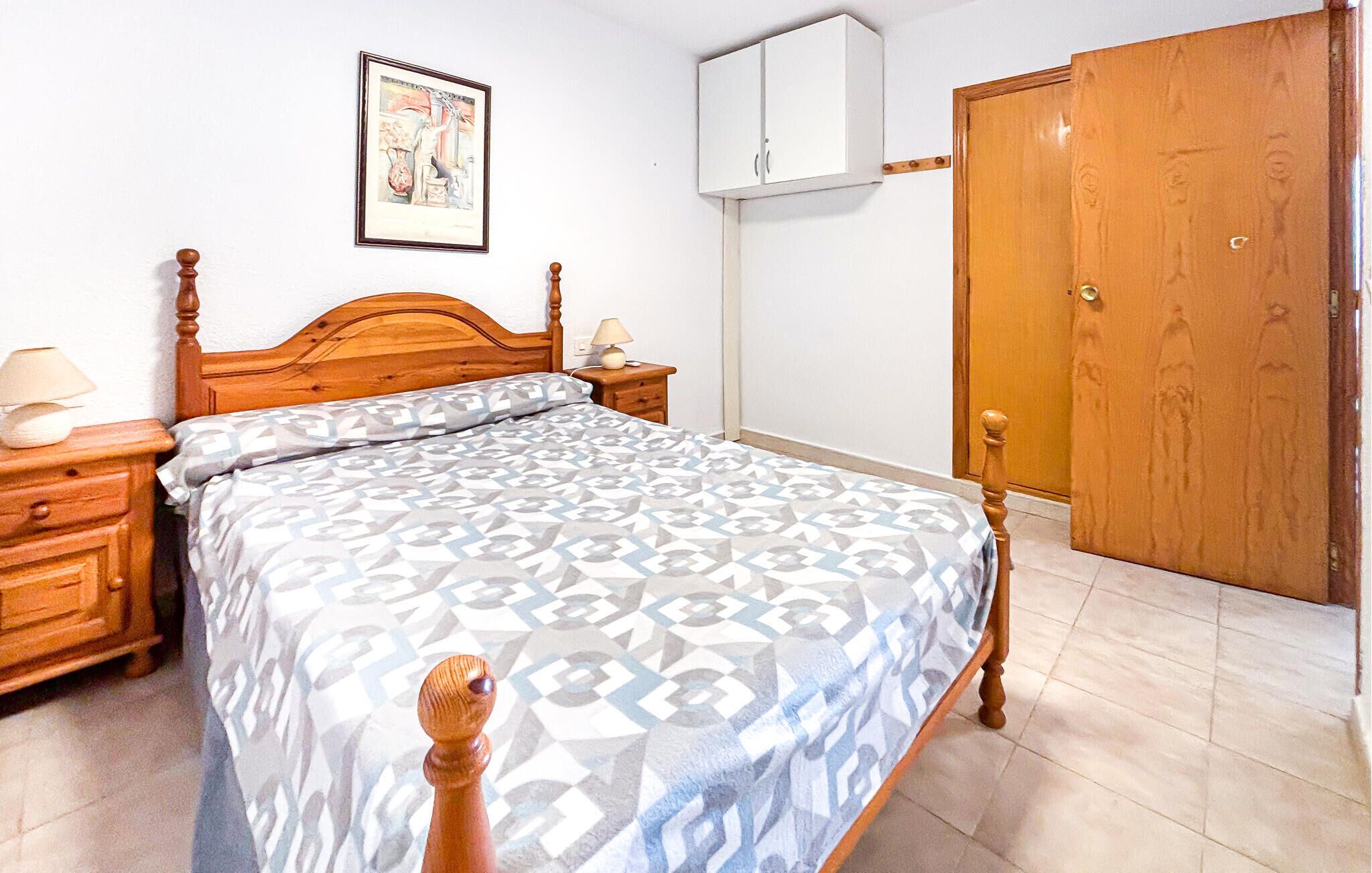 A bedroom with a wooden bed, a nightstand, a bedside table with a lamp, a framed picture on the wall, a white wardrobe, and a wooden door.