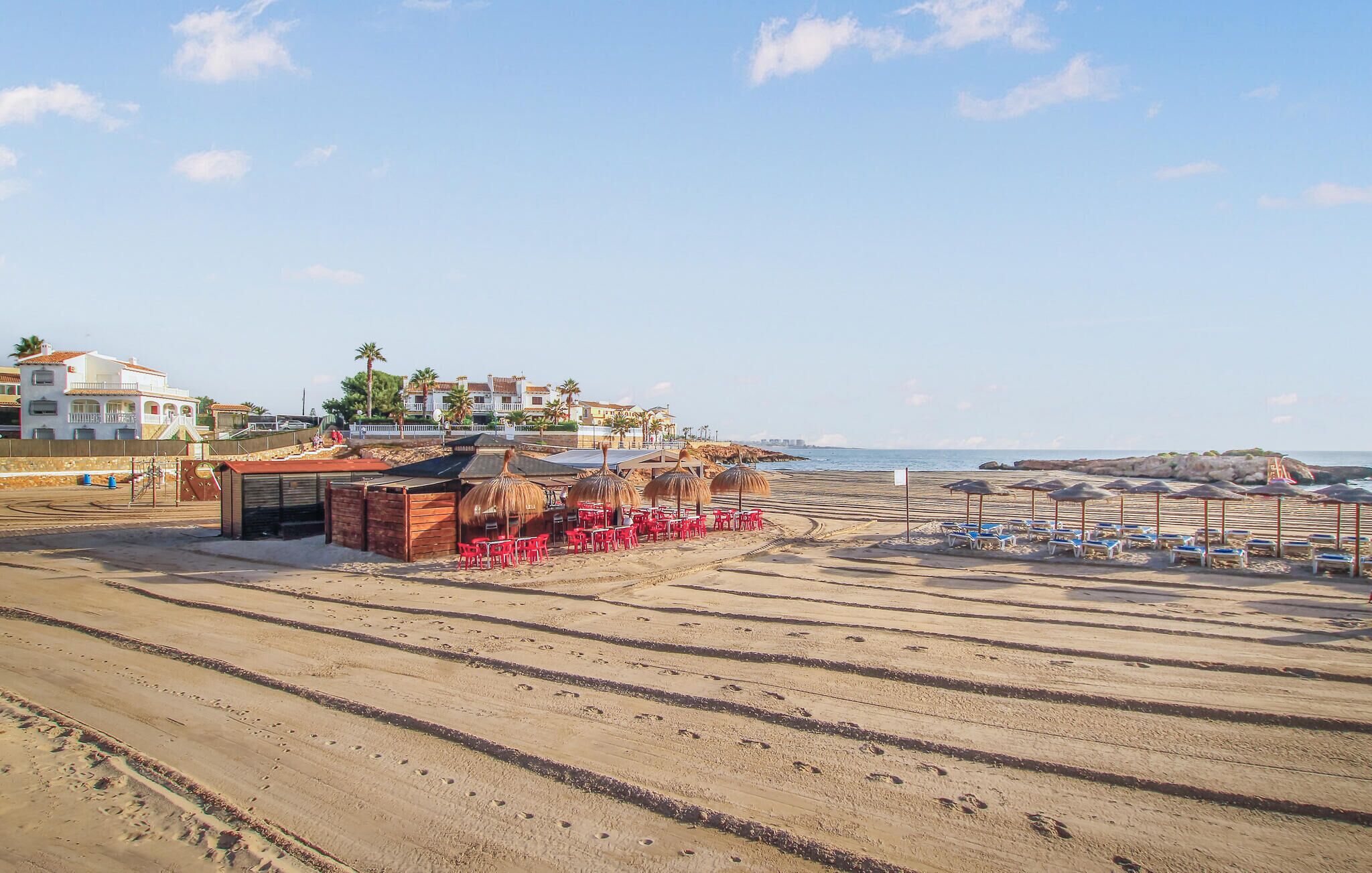 Ein Hotel direkt am Strand mit sandigem Ufer, roten Liegestühlen und Sonnenschirmen.