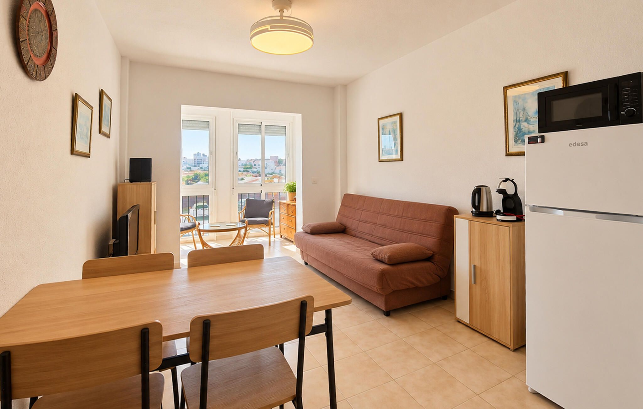 A compact living space with a dining table, chairs, a sofa, a kitchenette, and a view through large windows.