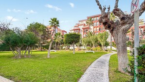 Um jardim bem cuidado com um caminho pavimentado, cercado por diversas árvores e arbustos, que leva a um edifício com fachada vermelha e várias varandas.