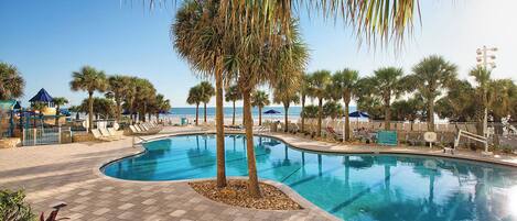 A swimming pool with a curved design, surrounded by palm trees and lounge chairs, with a beach and ocean view in the background.
