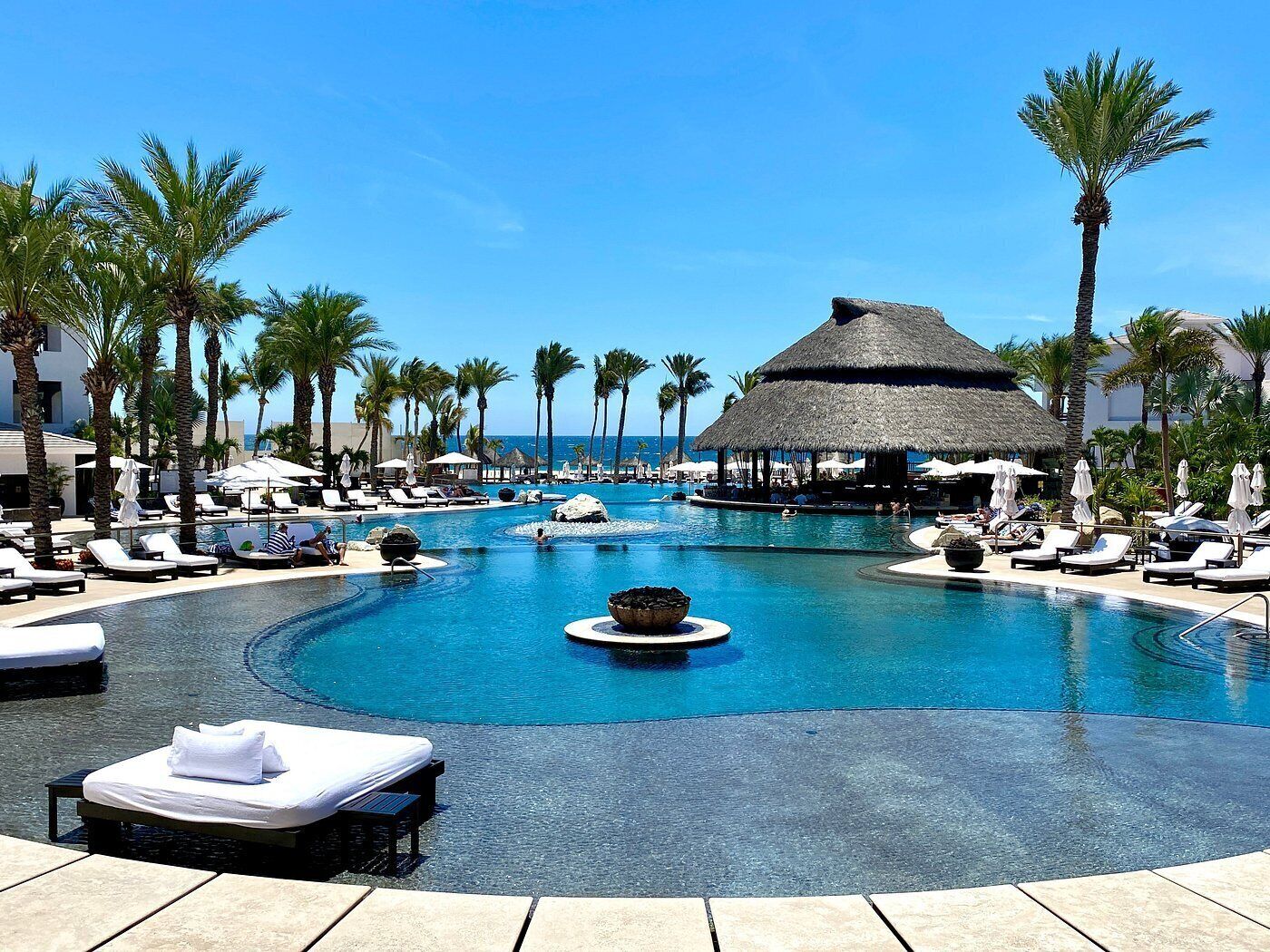 Piscina de resort con sillones de descanso, palmeras y una estructura de techo de paja.