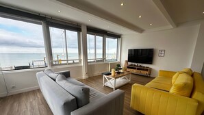 A modern living room with a large sofa, a yellow armchair, a coffee table, and a TV on a wooden cabinet. Large windows offer a view of the sea. - Captain YOO - The panorama / horizon, fabulous sea view (Fécamp)