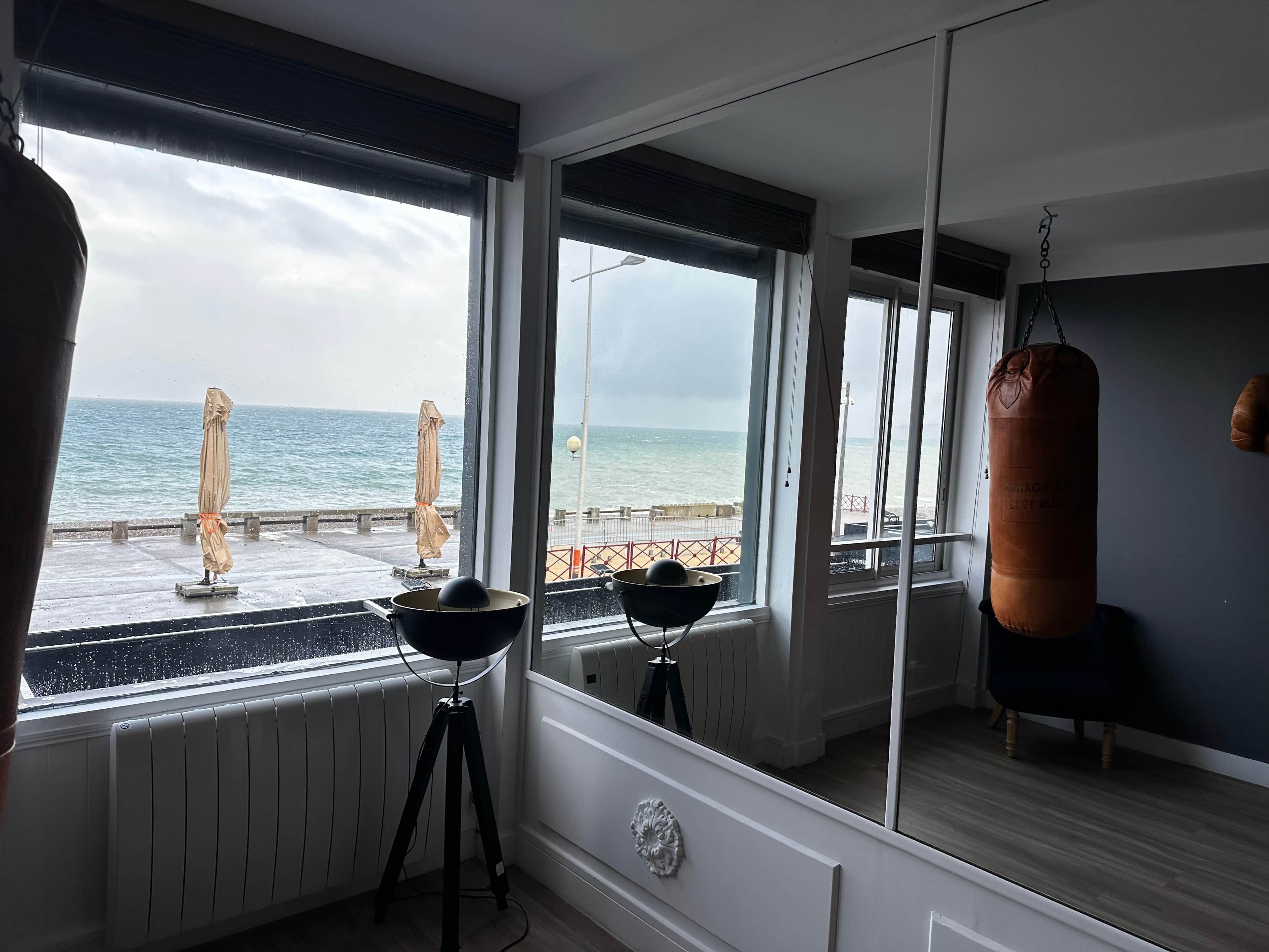 Een kamer met een groot raam dat uitkijkt op een steiger en de zee, een bokszak en een statief met een lamp.