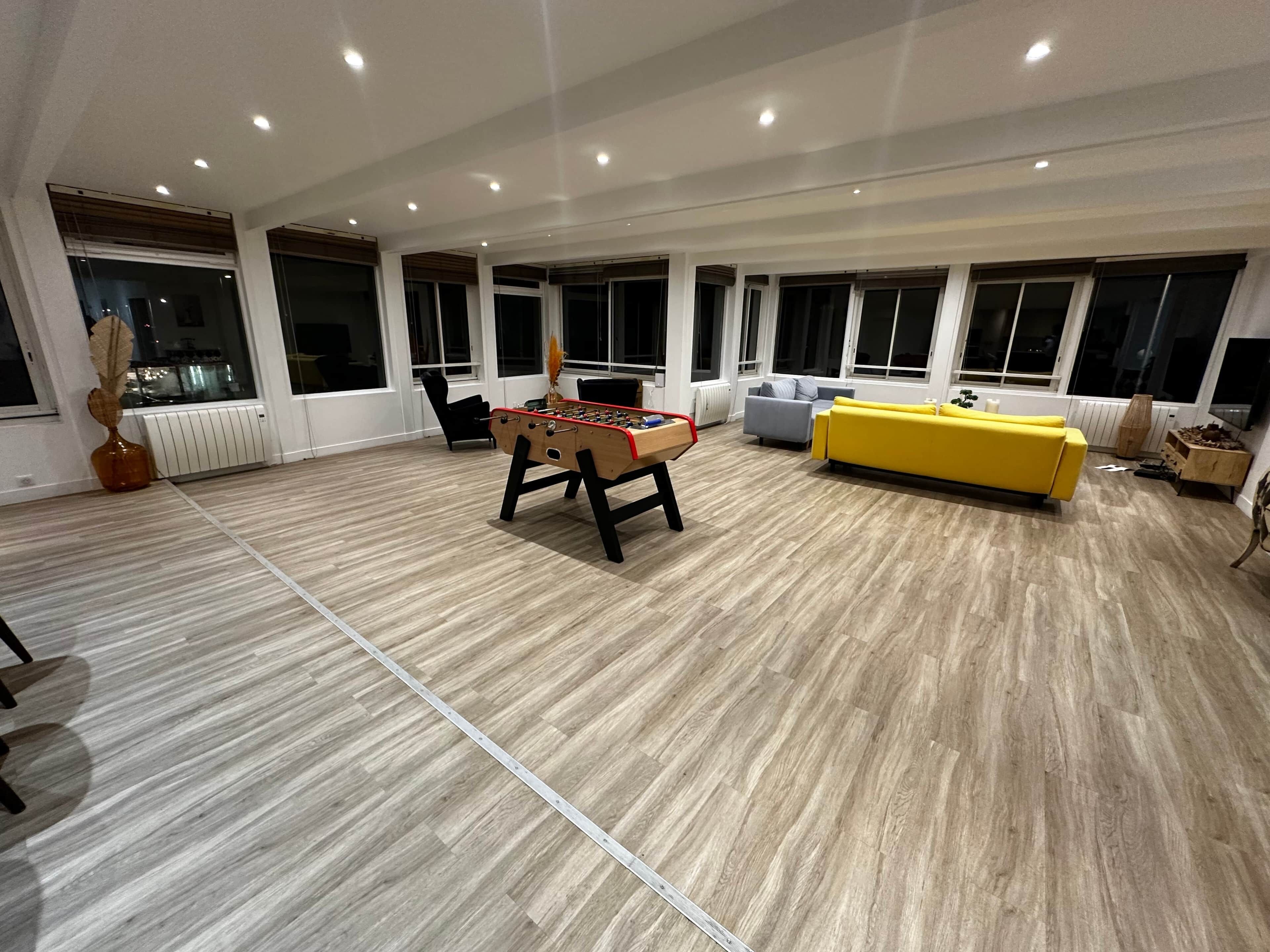 Een ruime kamer met een tafelvoetbalspel, een gele bank en grote ramen.