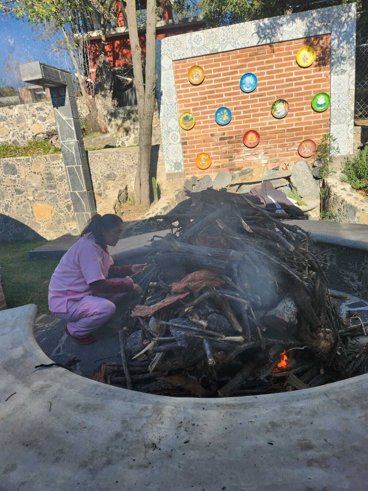 Uma pessoa cuidando de uma fogueira, com um muro de tijolos e pratos coloridos ao fundo.