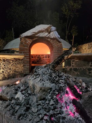 Un horno de ladrillo exterior con estructura abovedada, rodeado por un muro de piedra y una pila de carbón.