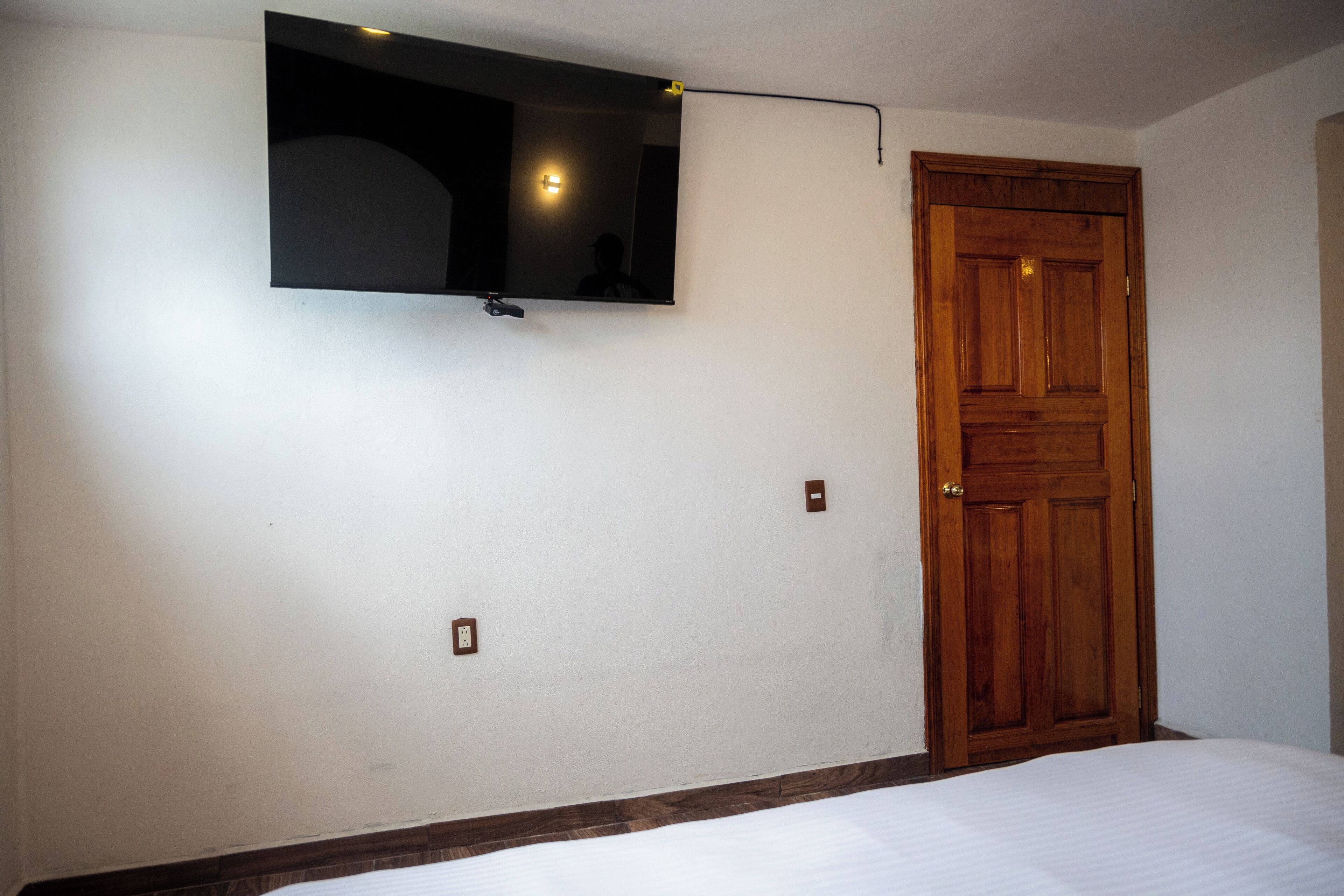 Een slaapkamer met een bed, een houten deur, een tv aan de muur en een lichtschakelaar.