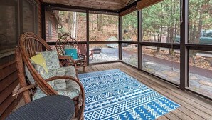 Eine überdachte Veranda mit Korbstühlen und einem gemusterten Teppich, große Fenster mit Blick auf ein bewaldetes Gebiet.