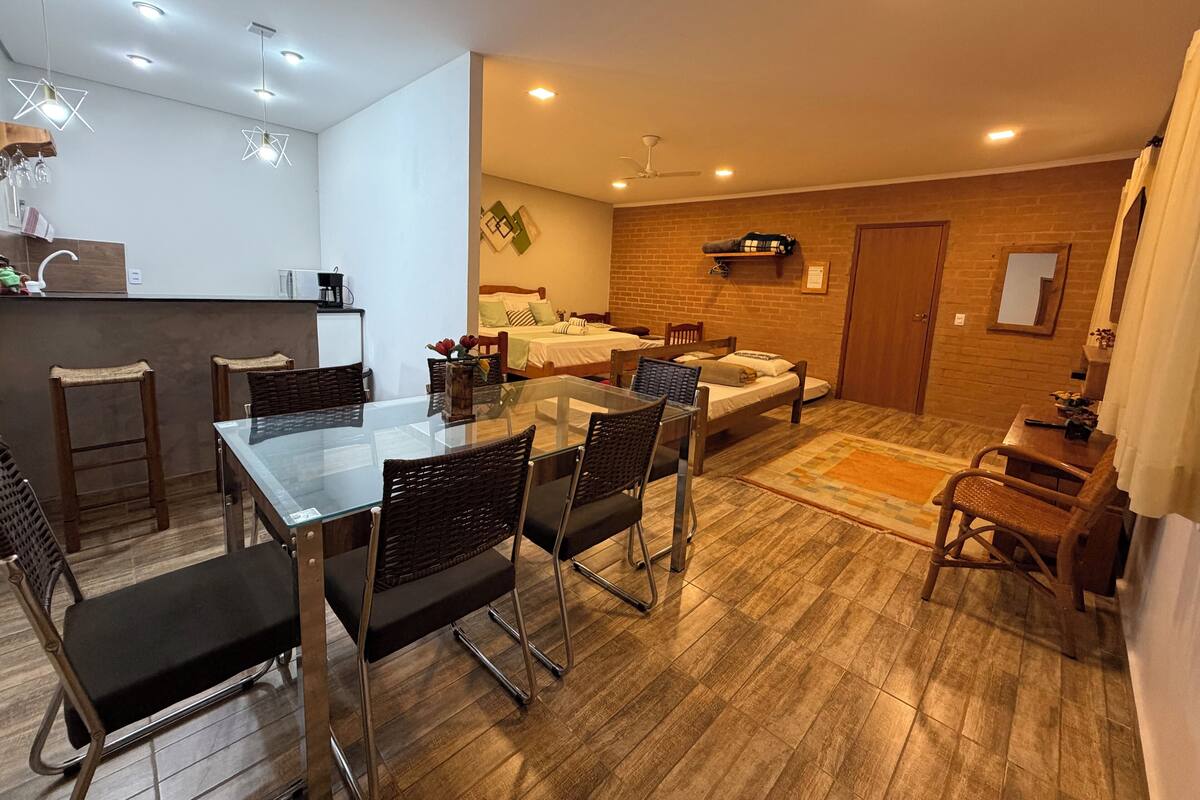 A modern hotel room with a glass dining table, wicker chairs, a sofa, and a bed.