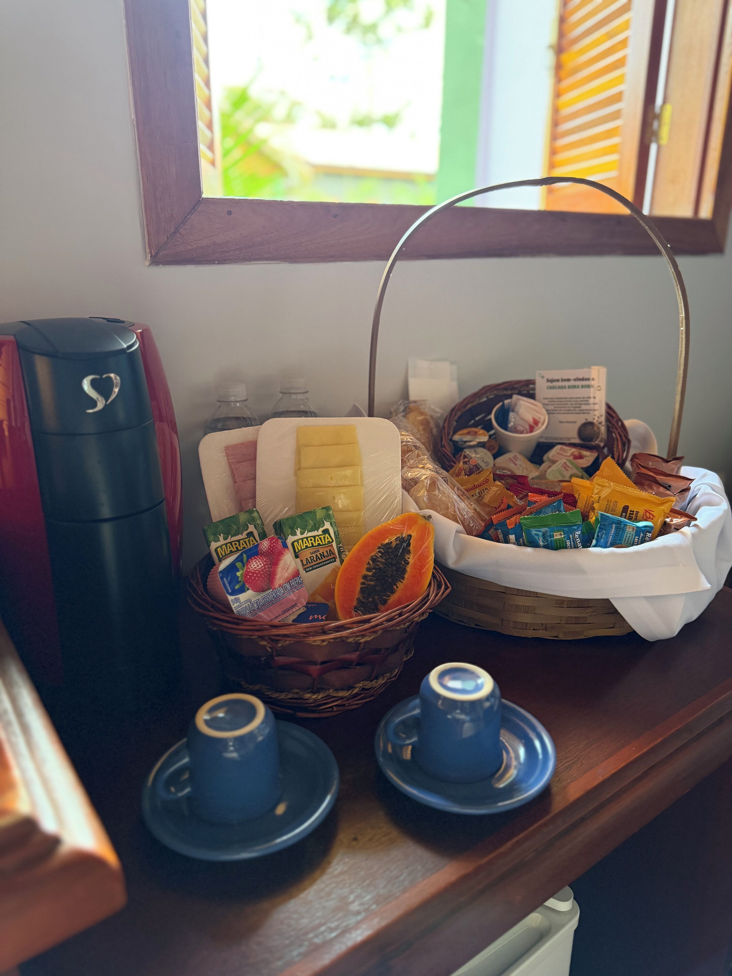 A breakfast setup with a coffee maker, a basket of snacks, and two cups on a wooden table.