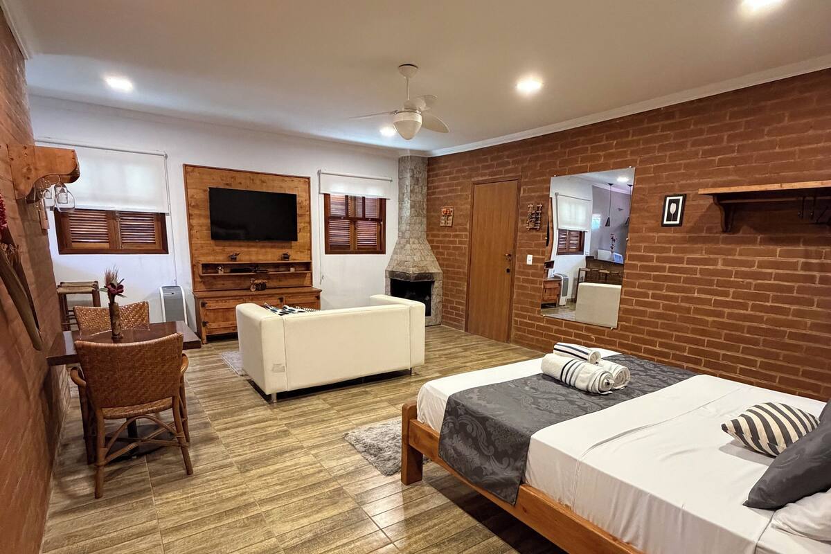 A modern hotel room with a wooden floor, a bed with a grey and white bedspread, a white sofa, a wooden coffee table with a vase of flowers, a television mounted on the wall, and a brick accent wall.