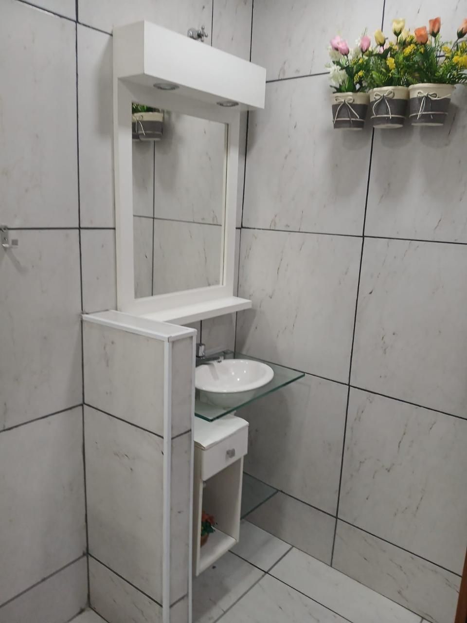 Een badkamer met een witte wastafel, een glazen wasbak en een spiegel. Boven de wasbak hangen drie bloempotten aan de muur.