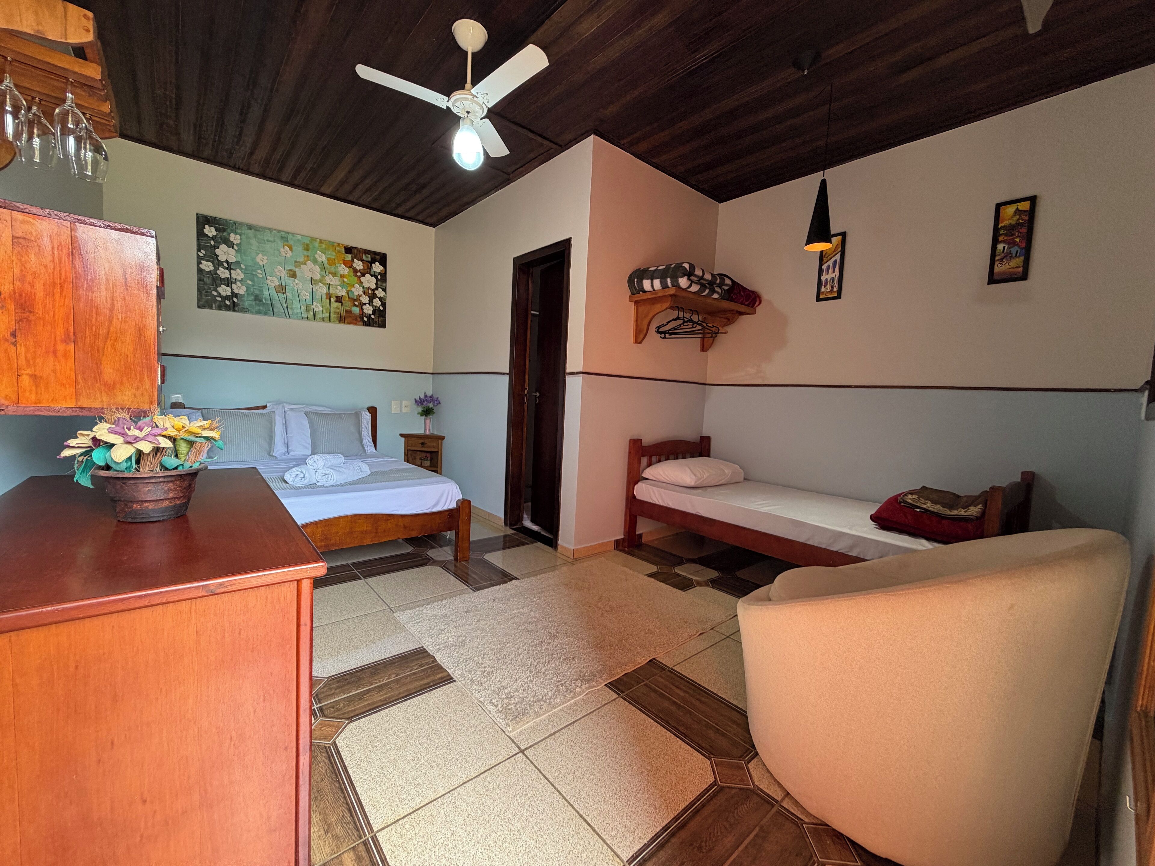 A bedroom with a wooden bed, a smaller bed, a chair, a desk with a vase of flowers, a ceiling fan, and a painting on the wall.