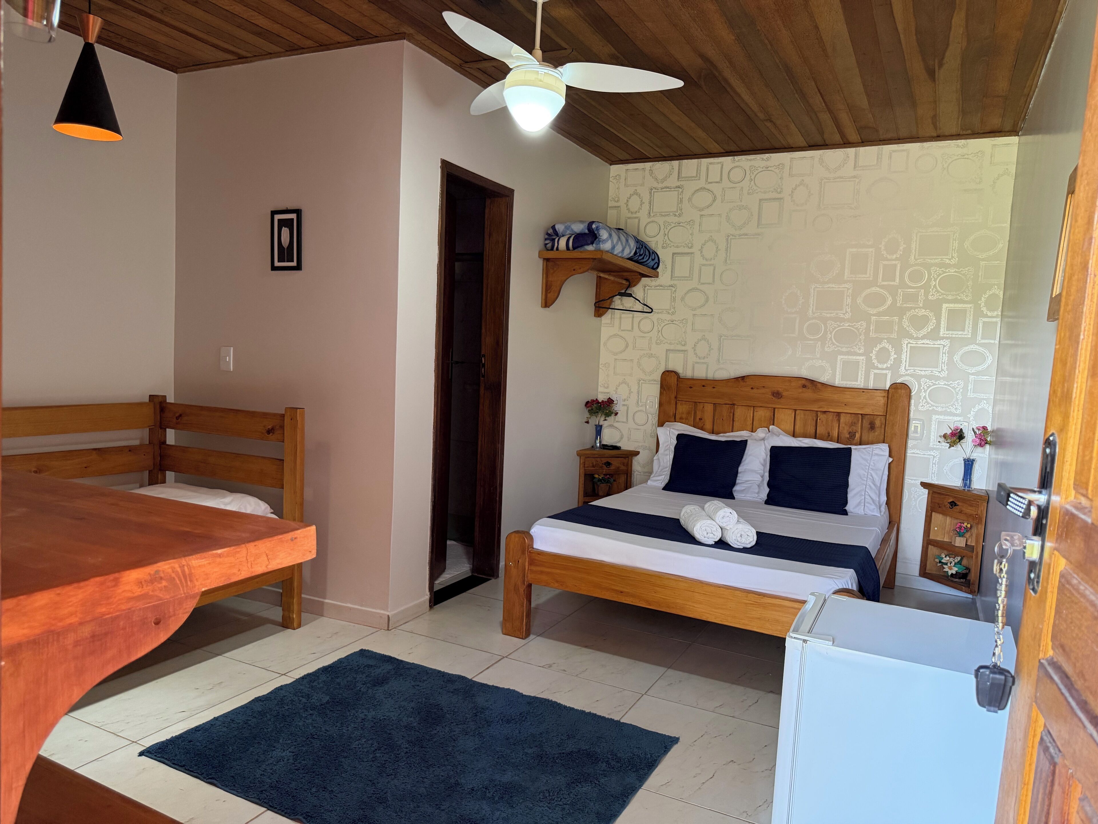 A bedroom with a wooden bed, a small bedside table, a nightstand, a ceiling fan, and a wall-mounted shelf.