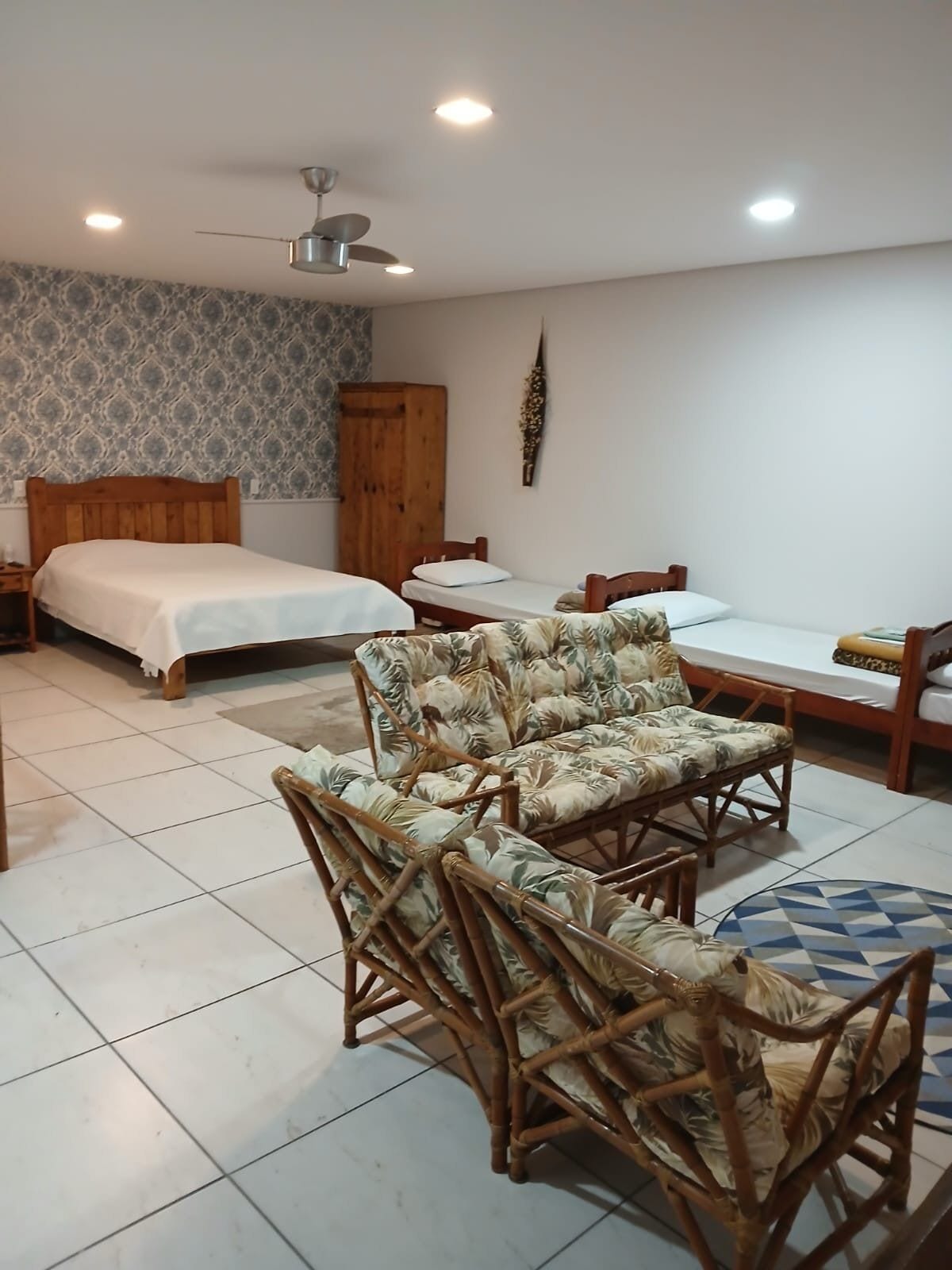Una habitación con dos camas, un armario de madera, ventilador de techo y un rincón de descanso con sillas de mimbre.