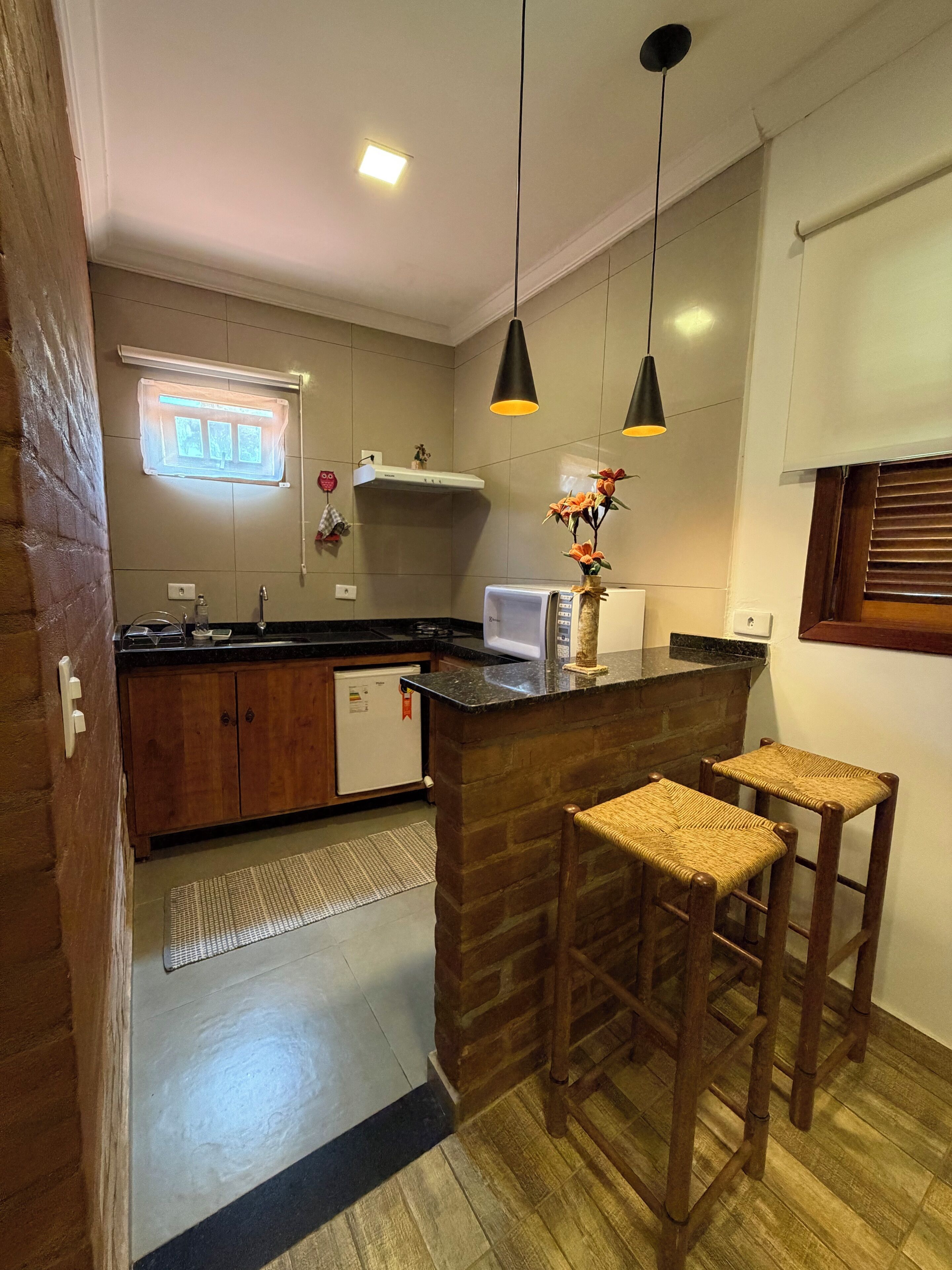 A kitchen with wooden cabinets, a refrigerator, a microwave, and a sink. There are two bar stools and a window with blinds.