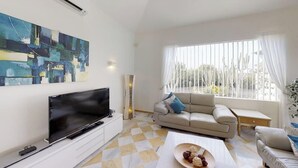 Un salon moderne avec un téléviseur à écran plat, un canapé beige, une table basse sur laquelle se trouve un panier de fruits, et une grande fenêtre ornée de rideaux blancs.
