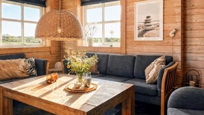 Interior acolhedor de uma cabana com uma mesa de jantar de madeira, uma luminária pendente de palha, um sofá e um quadro na parede.
