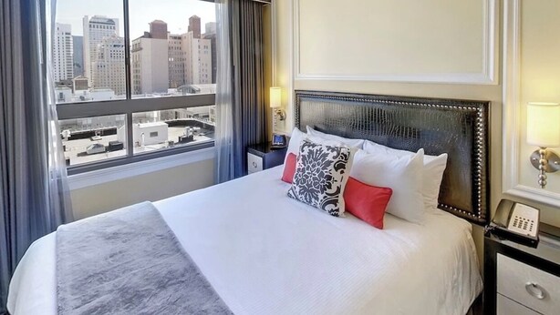 Habitación de hotel con una cama grande, vistas a la ciudad, una mesita de noche con lámpara y un aparador.
