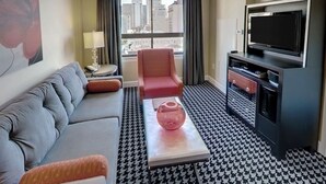 Habitación de hotel con un sofá gris, un sillón rojo, suelo estampado de pata de gallo, un televisor y una ventana con vistas a la ciudad.