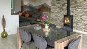 A modern living room with a glass dining table, grey upholstered chairs, a sofa with cushions, a decorative vase with flowers, a fireplace, and a stone wall. - 3 bedroom gorgeous home in Parigné (Parigné)