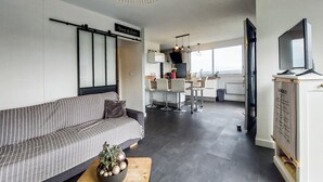 Uma sala de estar moderna com um sofá listrado, uma mesa de centro com uma planta e uma área de refeições, com uma cozinha ao fundo.