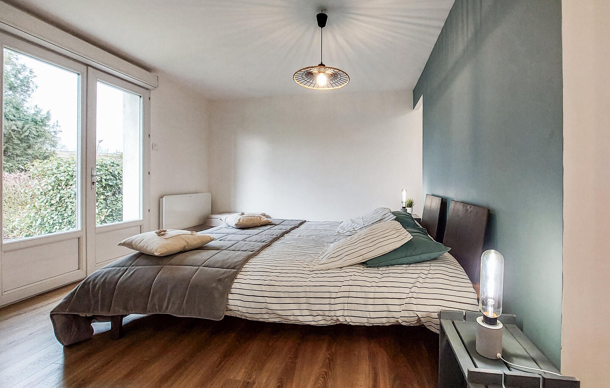Et soveværelse med et stort vindue, en seng med stribet sengetøj, en sengetavlelampe og en hængende lampe.