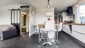 Uma cozinha moderna com armários brancos, uma sala de jantar com mesa de madeira e cadeiras brancas, e uma sala de estar com sofá e televisão.