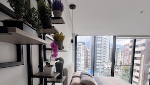 A modern bedroom with a bed, built-in shelves with plants, a bedside table, and a view of tall buildings through large windows. - Deluxe green suite/ La Carolina (Quito)