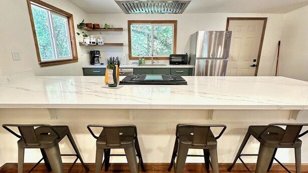 A modern kitchen with a large island, bar stools, and a stainless steel refrigerator. - Large Cabin on Ave of Giants with private Eel River access (Miranda)