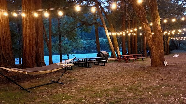 Eine Hängematte, Picknicktische und Lichterketten in einer bewaldeten Gegend mit einem See.