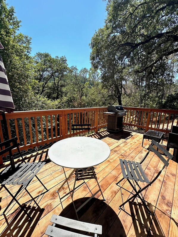 A wooden deck with a round table and metal chairs, surrounded by a wooden fence, with trees in the background.