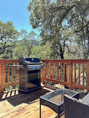 A barbecue grill on a wooden deck with a glass table and wicker chairs.
