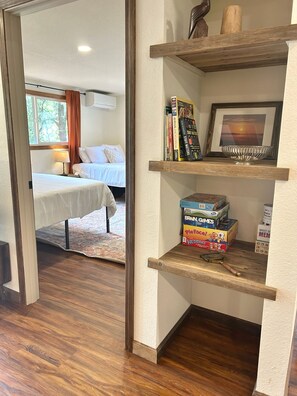 Ein Hotelzimmer mit Holzboden, einem Bett mit weißer Bettwäsche, einem Nachttisch mit Lampe, einem Bücherregal mit Büchern und Spielen und einem gerahmten Bild an der Wand.
