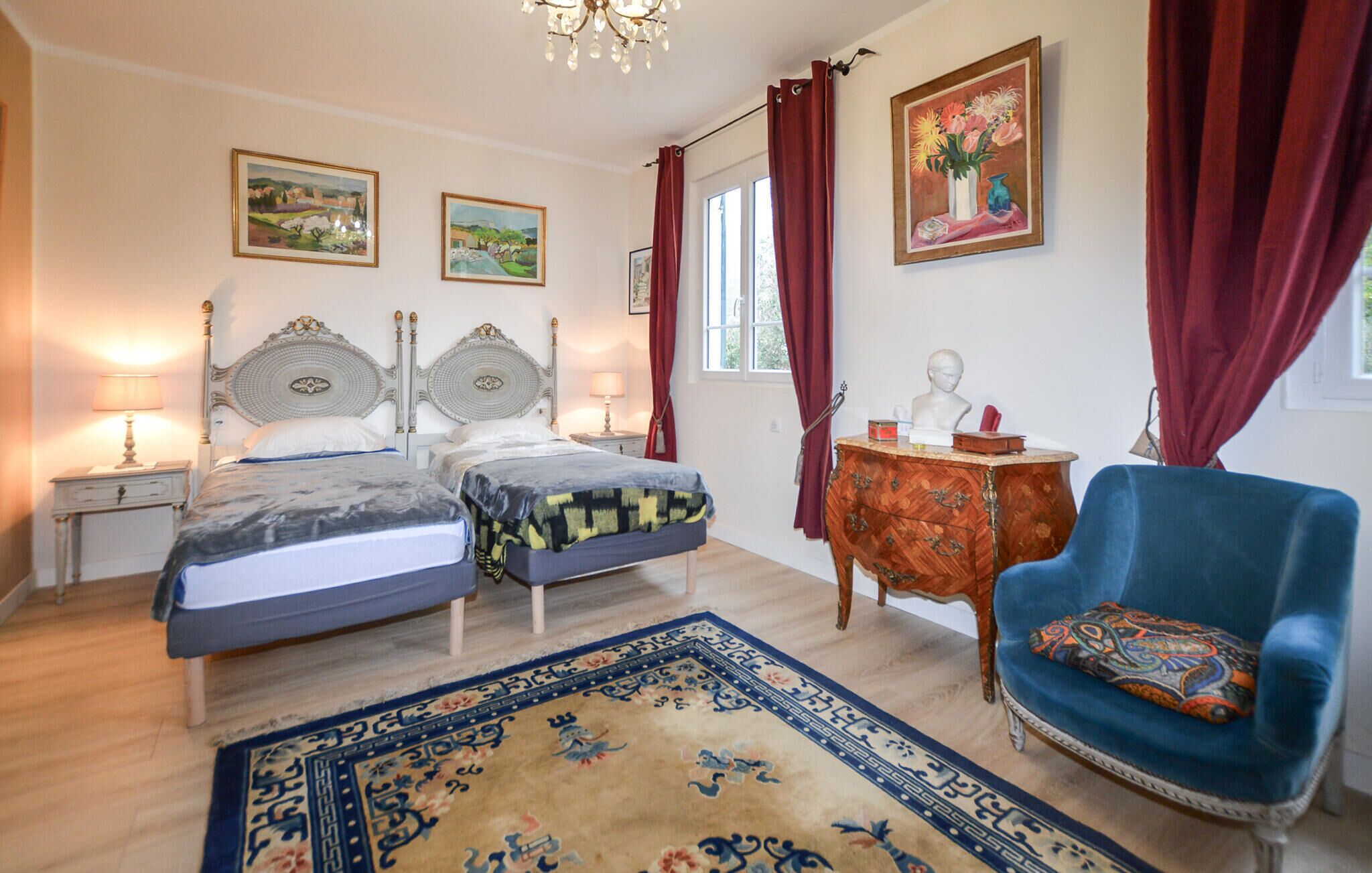 Een slaapkamer met twee bedden, een blauwe fauteuil, een houten dressoir en een groot vloerkleed.