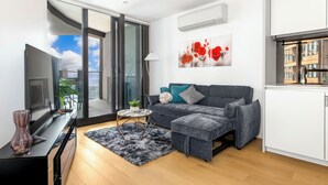 Un salon moderne avec un canapé gris, une petite table basse ronde, un meuble TV noir et une grande fenêtre donnant sur la ville.