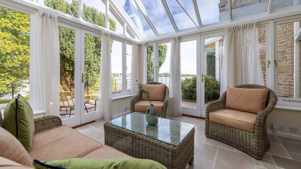 Ein Wintergarten mit Korbmöbeln, einem Glastisch und Blick auf das Grün draußen.