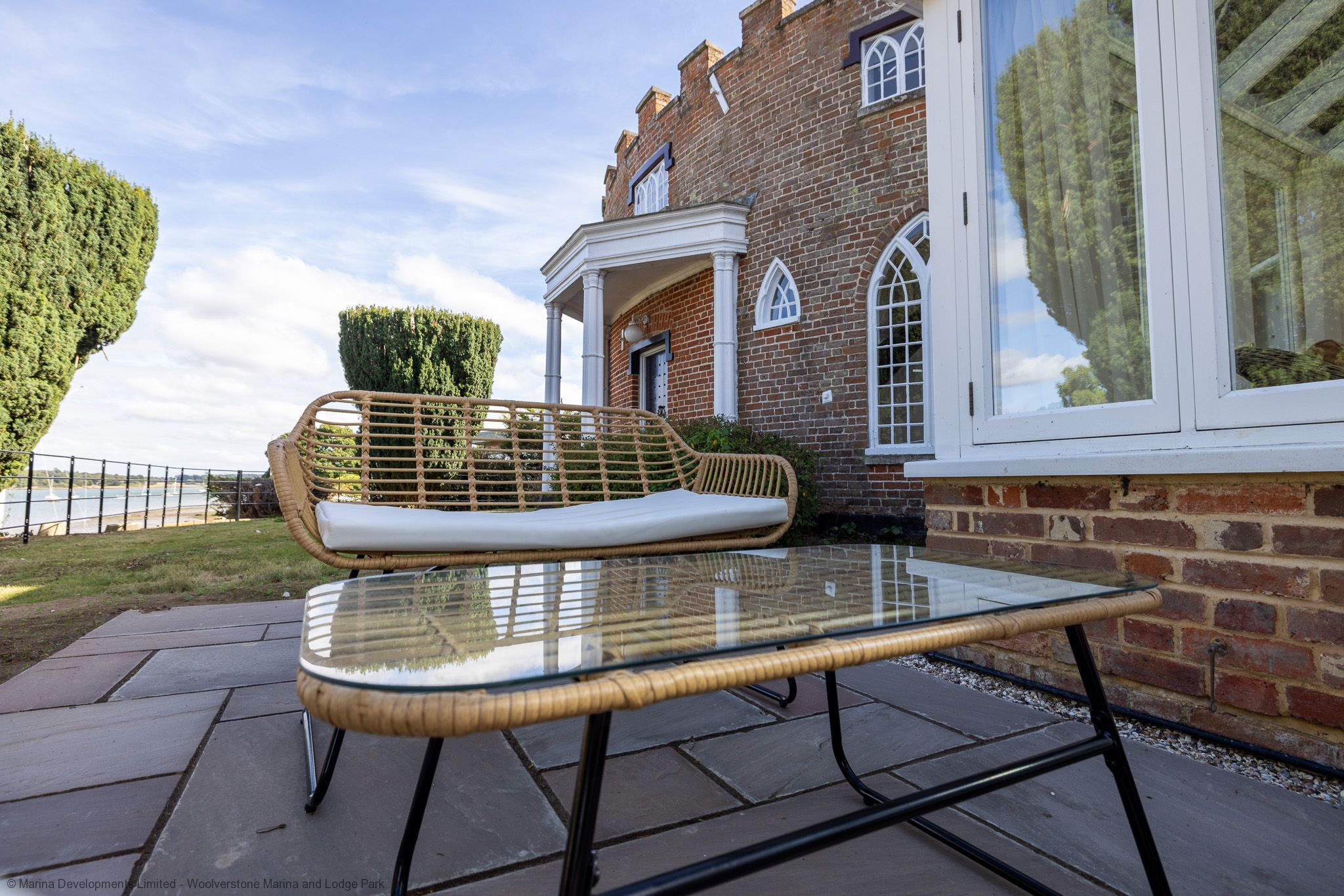A glass-top table with a wicker chair and a cushion, positioned on a stone patio in front of a brick building with arched windows and a white porch.