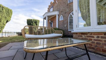 A glass-top table with a wicker chair and a cushion, positioned on a stone patio in front of a brick building with arched windows and a white porch.