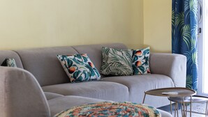 Een woonkamer met een grijze bank, twee gekleurde kussens, een gehaakte plaid en een klein rond tafeltje.