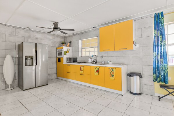A modern kitchen with a stainless steel refrigerator, a microwave, and a coffee maker. There is a ceiling fan and a window with curtains.