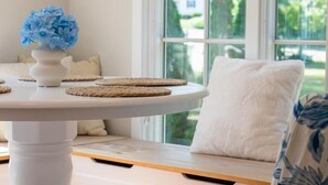 A white table with a vase of blue flowers, woven coasters, and a white cushion on a bench by a window. - Family Cottage with 4 King Beds  (Norwalk)