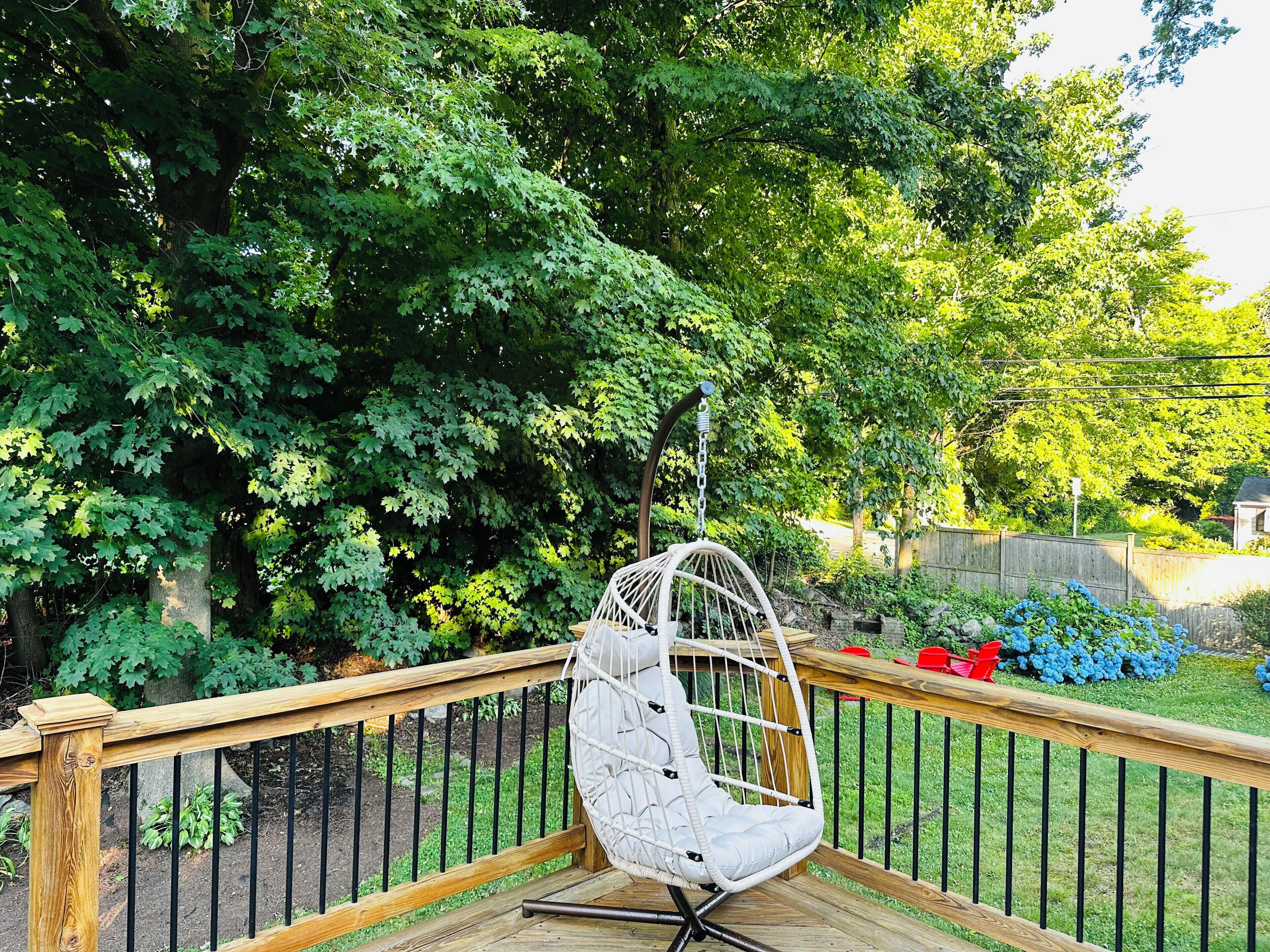 A wooden deck with a hanging chair, surrounded by lush greenery and a wooden fence.